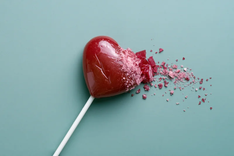 Broken red heart-shaped lollipop with shattered pieces on teal background