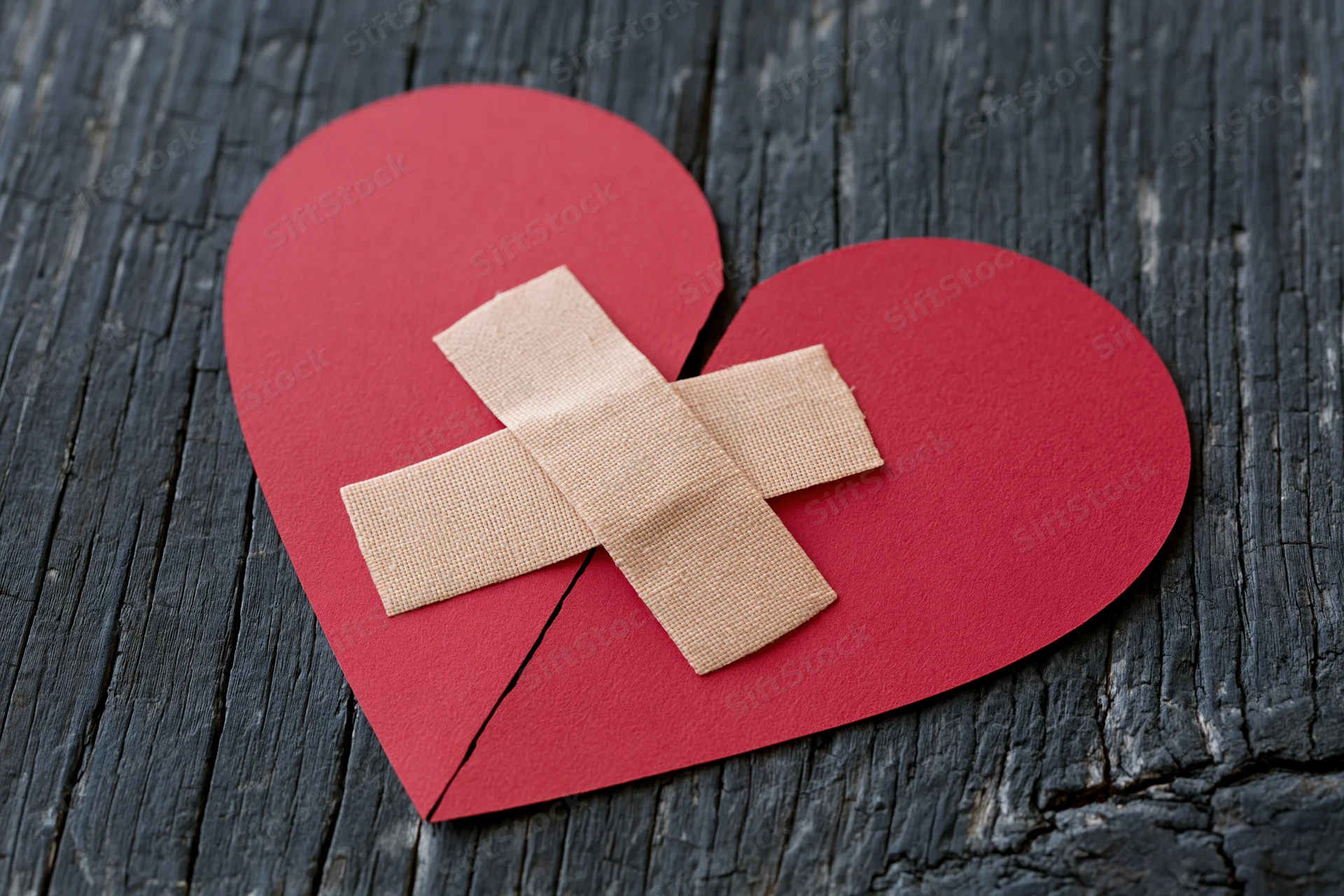 Red paper heart split down the middle with two adhesive bandages forming a cross on dark wood surface
