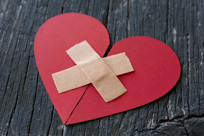 Red paper heart split down the middle with two adhesive bandages forming a cross on dark wood surface