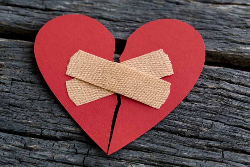 Red paper heart split down the middle with two adhesive bandages on weathered wood