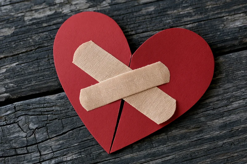 Red paper heart split down the middle with two bandages forming a cross on weathered wood