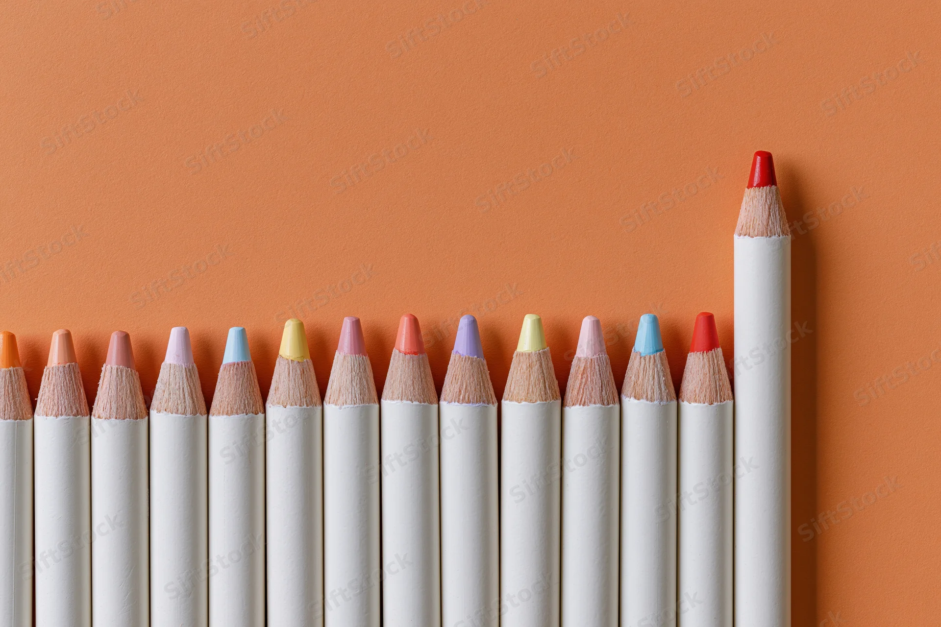 Row of white colored pencils with multicolored tips arranged in line on orange surface, one red pencil standing taller than others