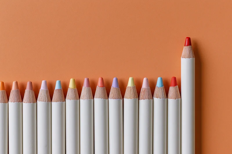 Row of white colored pencils with multicolored tips arranged in line on orange surface, one red pencil standing taller than others