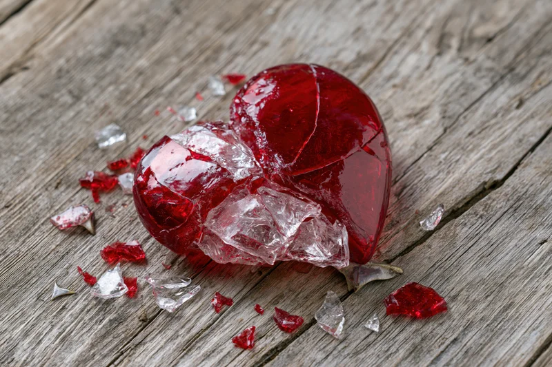 Shattered red glass heart resting on weathered wooden surface with scattered fragments