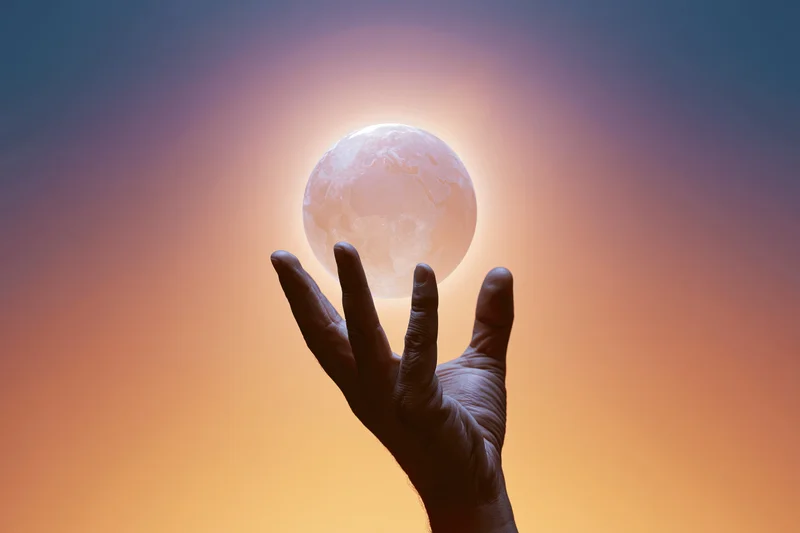 Silhouetted hand reaching upward toward glowing earth against sunset sky