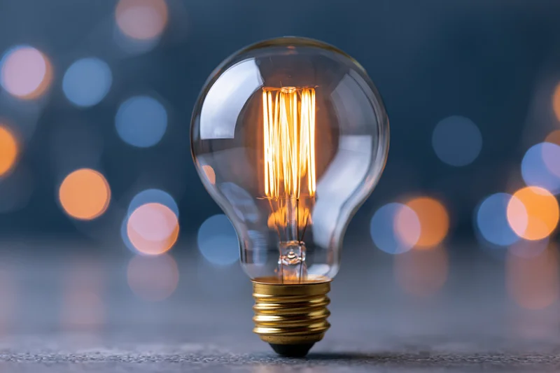 Glowing incandescent light bulb with visible filament standing on a textured surface against a blurred bokeh background