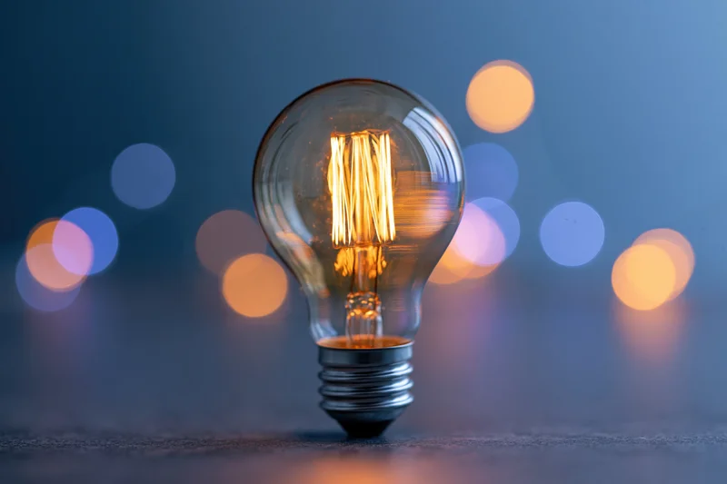 Glowing incandescent light bulb with visible filament standing on a dark surface against a blurred bokeh background