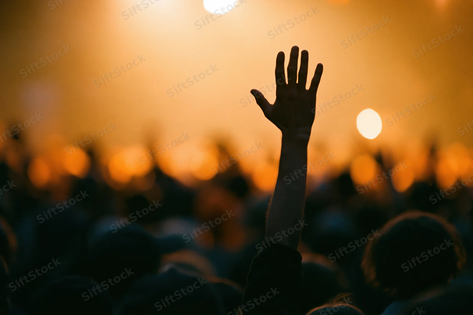Hand raised above a crowd of silhouetted figures at a concert under warm stage lights