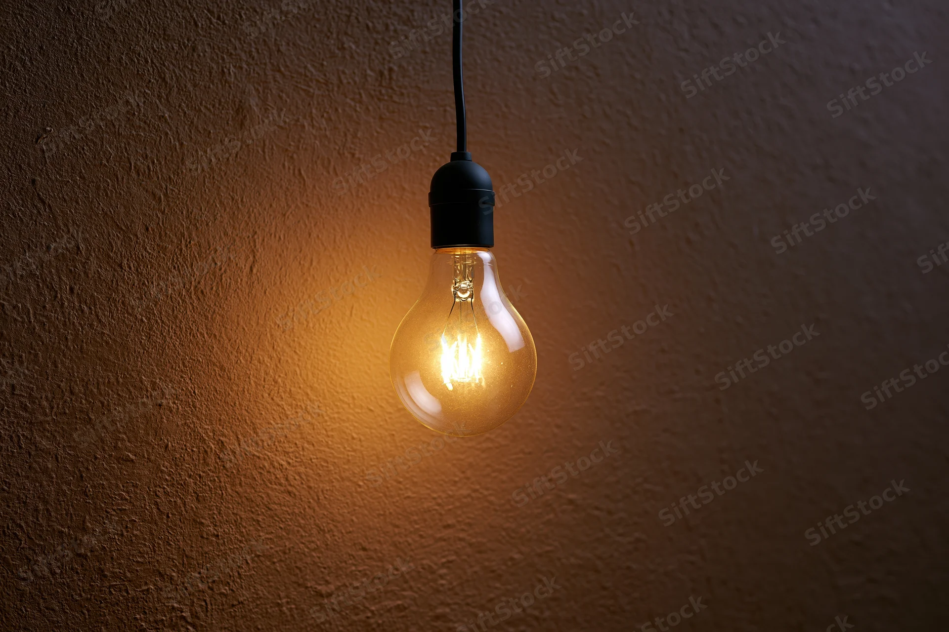 Illuminated incandescent lightbulb hanging from black wire against textured wall