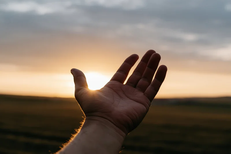 Open hand reaching upward toward the sunset over a field