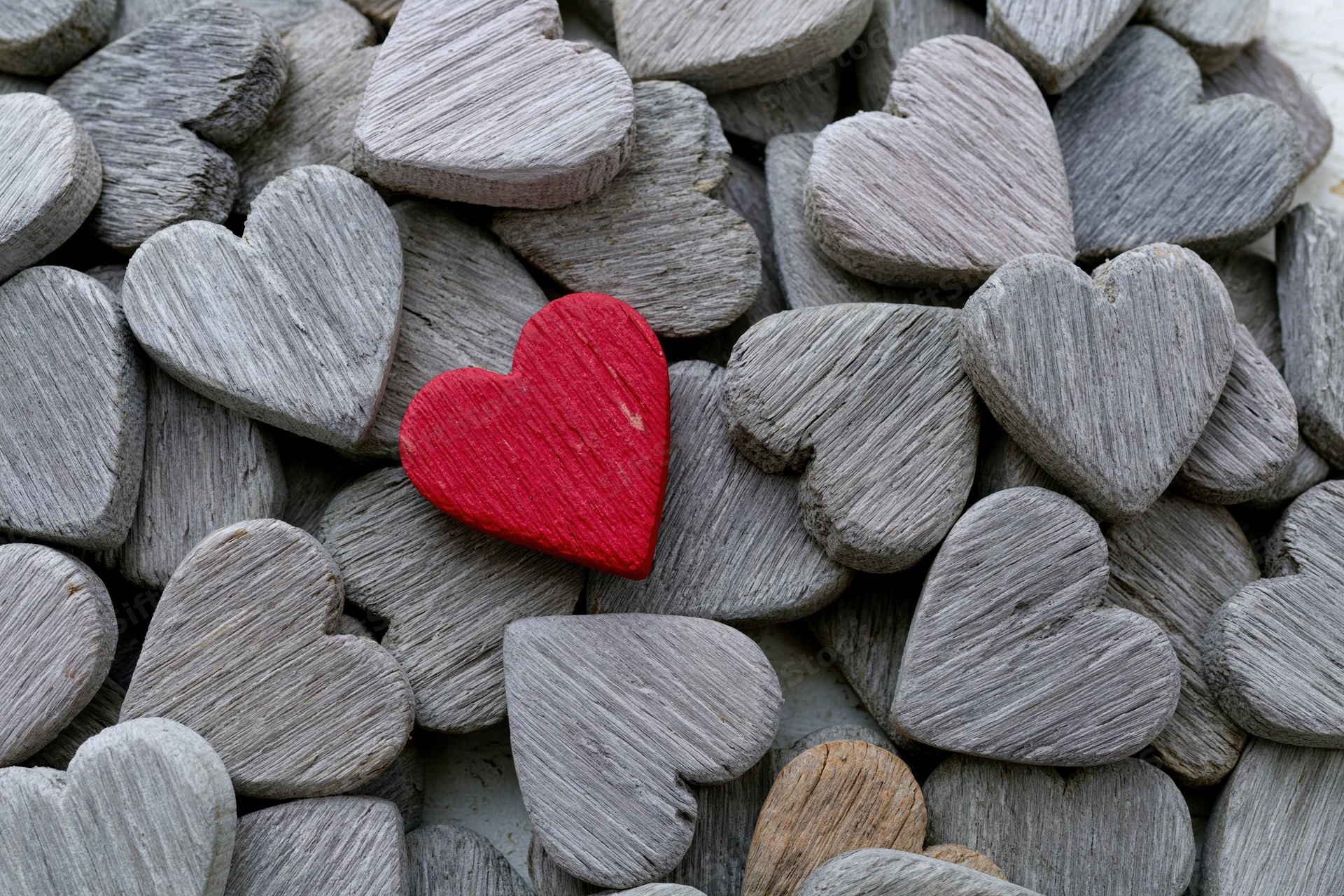 Red painted wooden heart resting among gray weathered wooden hearts in a pile