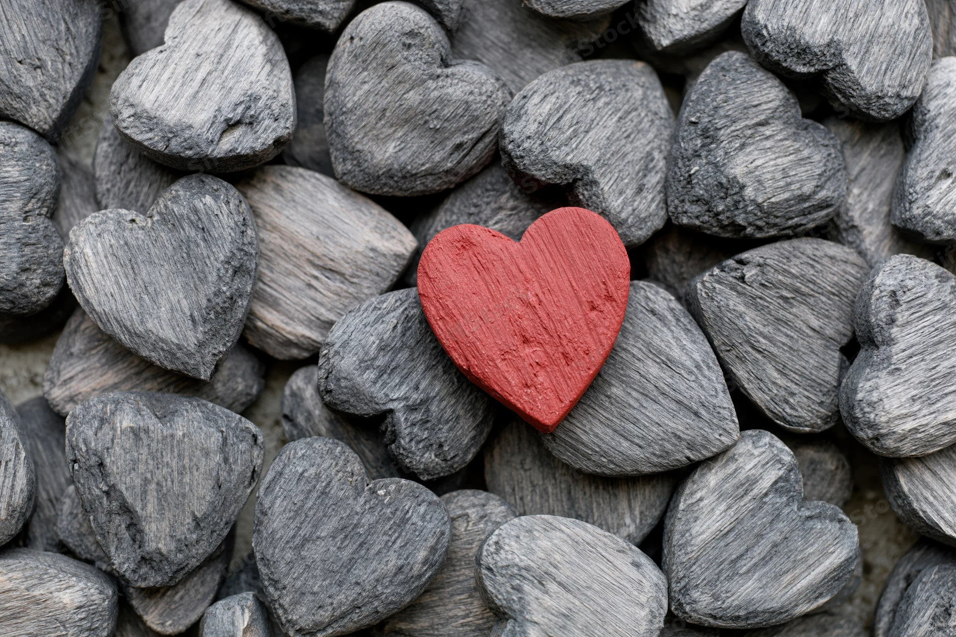 Red painted wooden heart resting among gray weathered stone hearts in a pile