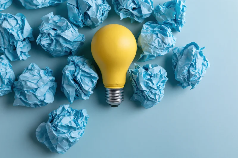 Yellow lightbulb surrounded by crumpled blue paper on a light blue surface