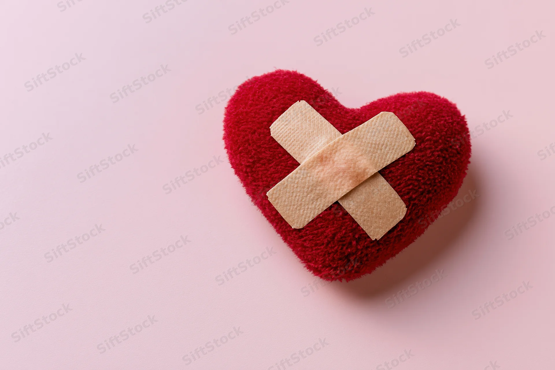Red plush heart with beige bandages in cross shape on pink surface