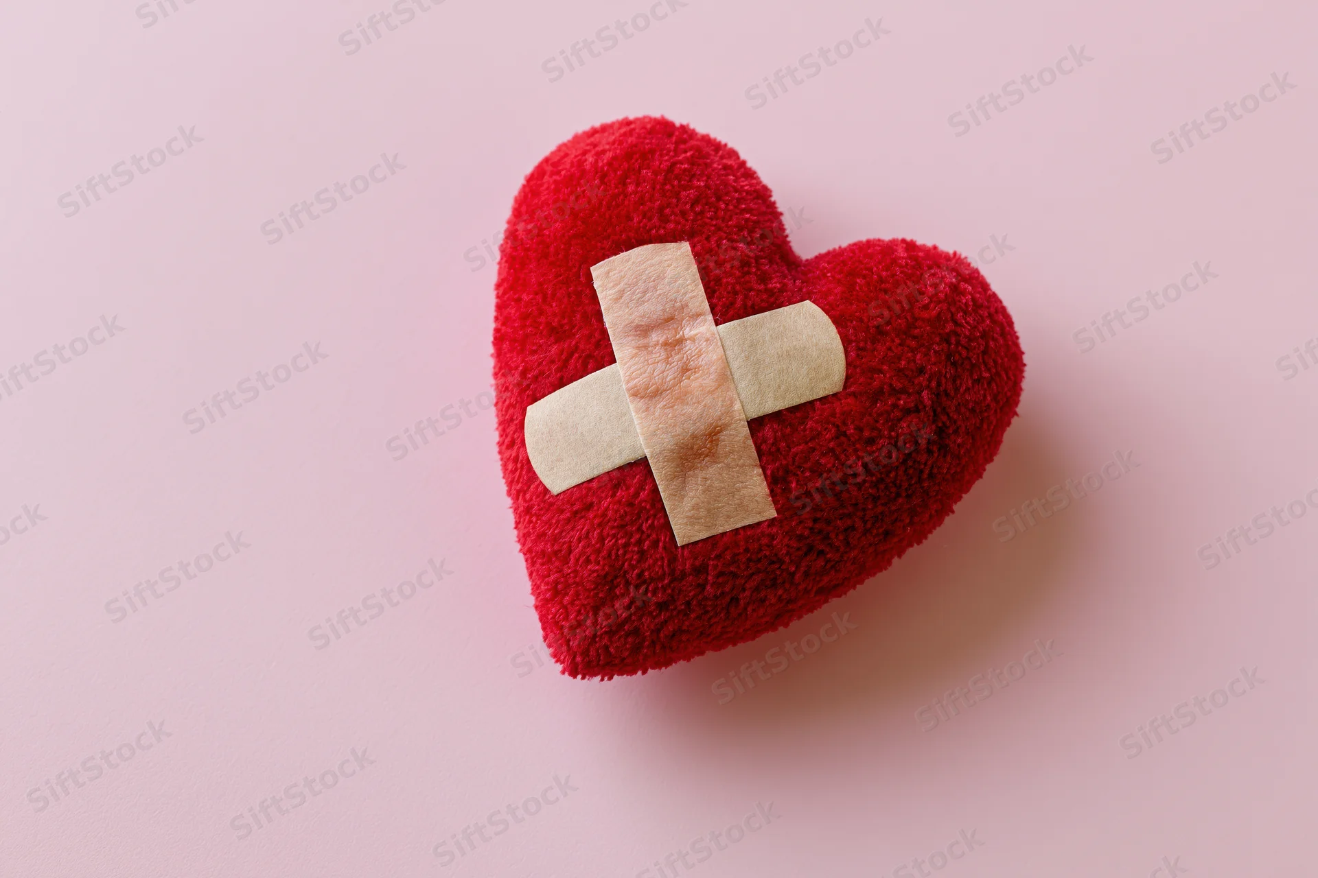 Red plush heart with beige bandages forming a cross on pink background