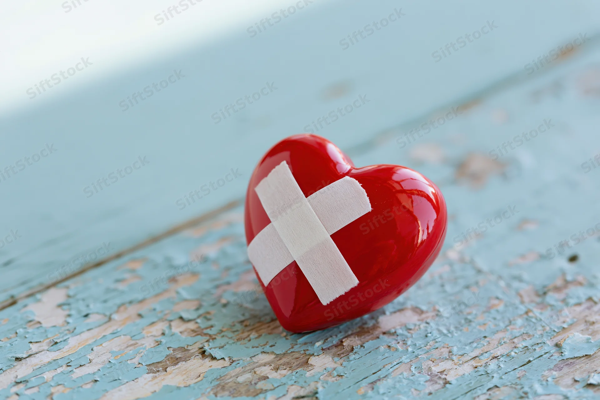 Glossy red ceramic heart with white bandage cross on peeling blue wooden surface