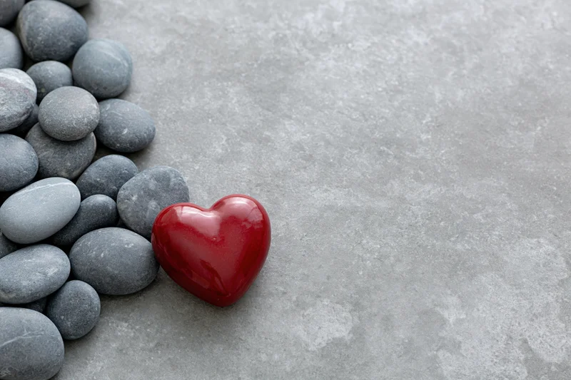 Glossy red ceramic heart resting among smooth gray pebbles on concrete surface