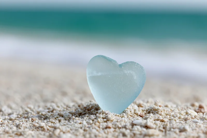 Heart-shaped sea glass resting on sandy beach with ocean in background