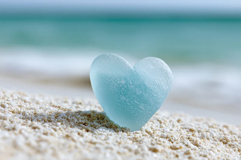 Heart-shaped sea glass resting on sandy beach with ocean in background