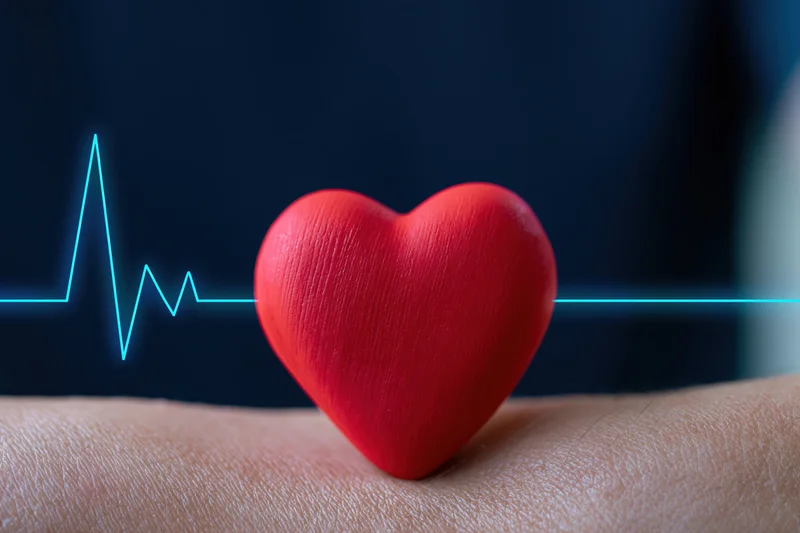 Red heart object resting on human wrist with digital heartbeat line in dark background