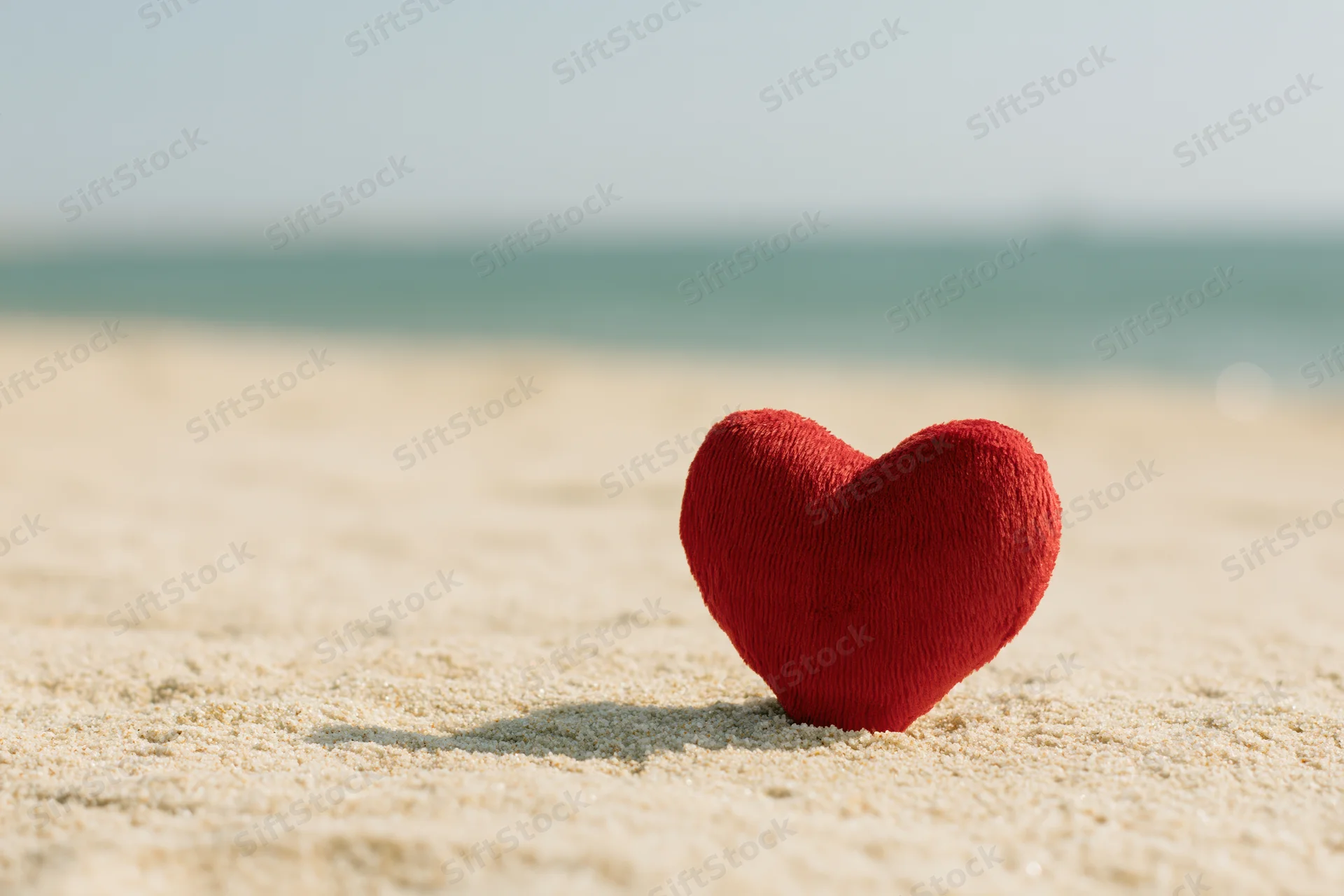 Red plush heart standing upright on sandy beach near ocean