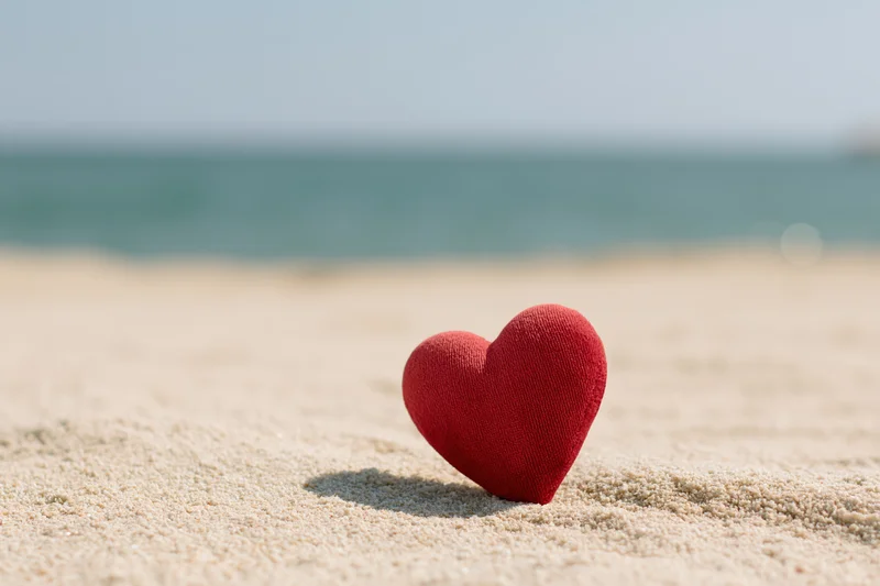 Red plush heart standing upright on sandy beach near ocean