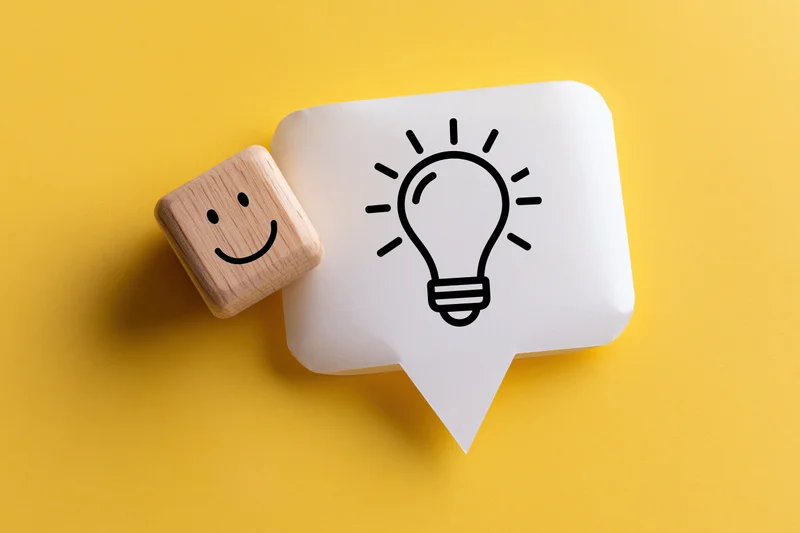Wooden cube with smiley face next to speech bubble containing lightbulb icon on yellow background