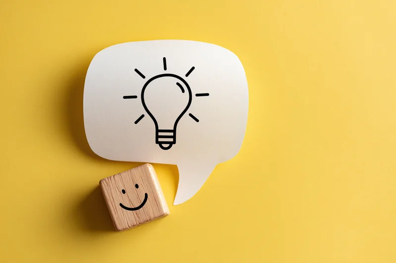 Wooden block with smiley face beside a speech bubble containing a lightbulb on yellow background