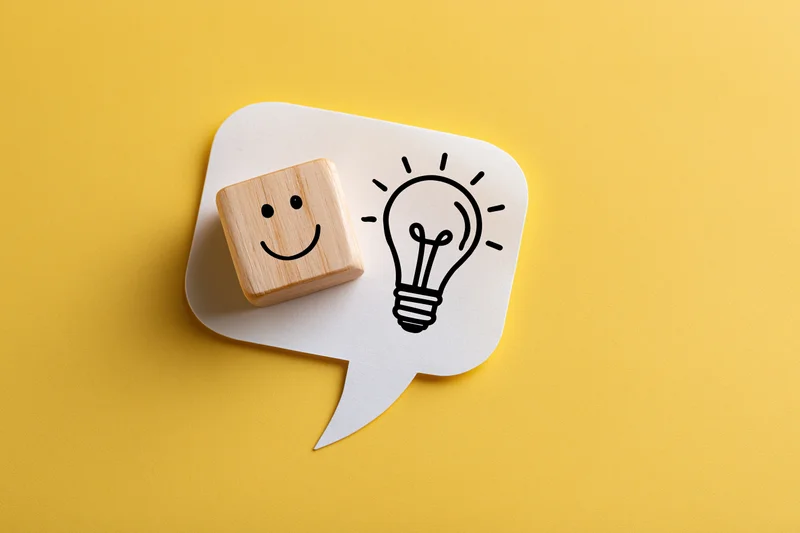 Wooden cube with smiley face and drawn lightbulb inside speech bubble on yellow background
