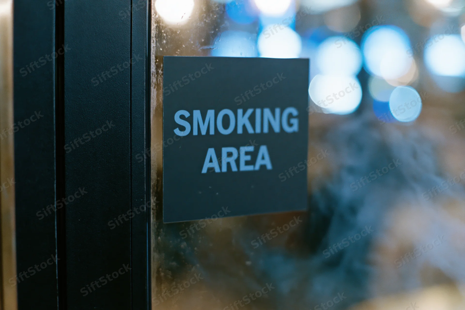 Black sign reading smoking area on glass door with smoke and blurred lights in background
