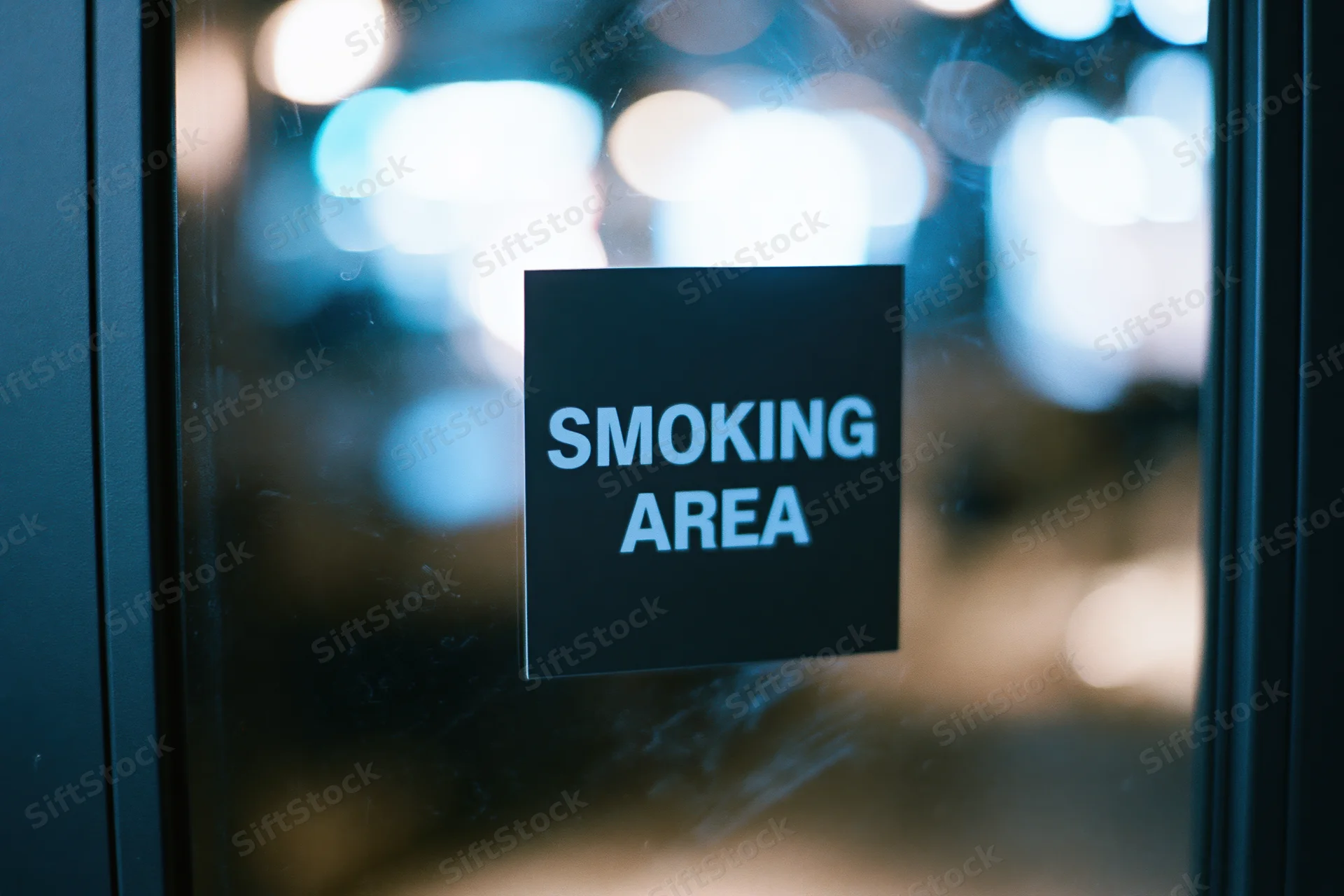 Black square sign reading smoking area on glass door with blurred city lights behind