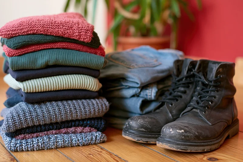 Stack of folded colorful knitwear and sweaters next to worn boots and jeans on wooden floor