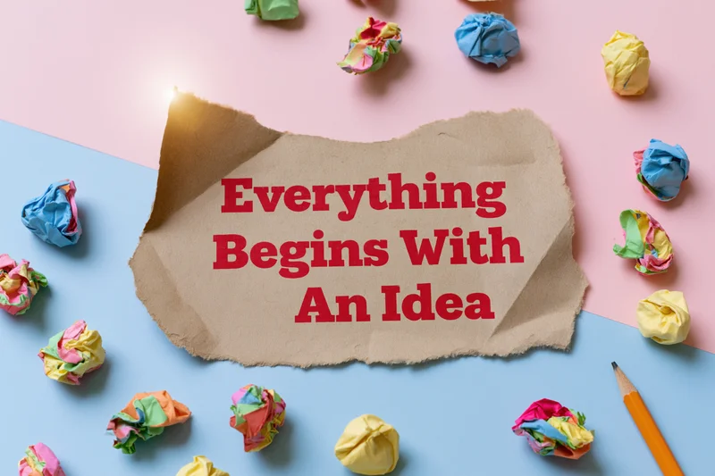 Torn kraft paper with bold red text reading everything begins with an idea surrounded by crumpled colorful papers and a pencil on a pink and blue surface