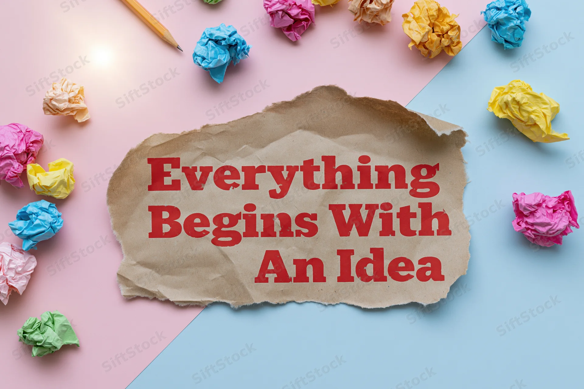 Torn kraft paper with red text reading everything begins with an idea surrounded by crumpled colorful papers and a pencil on pink and blue background