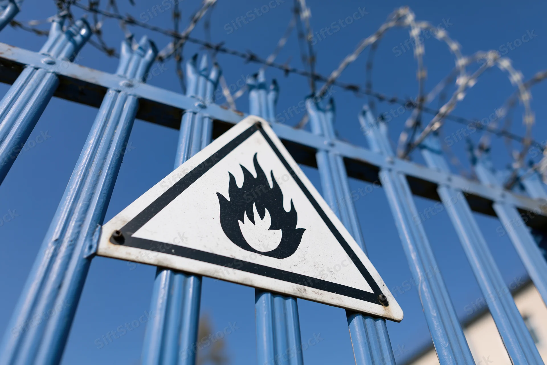 Triangular flammable warning sign mounted on blue metal fence with barbed wire against clear blue sky