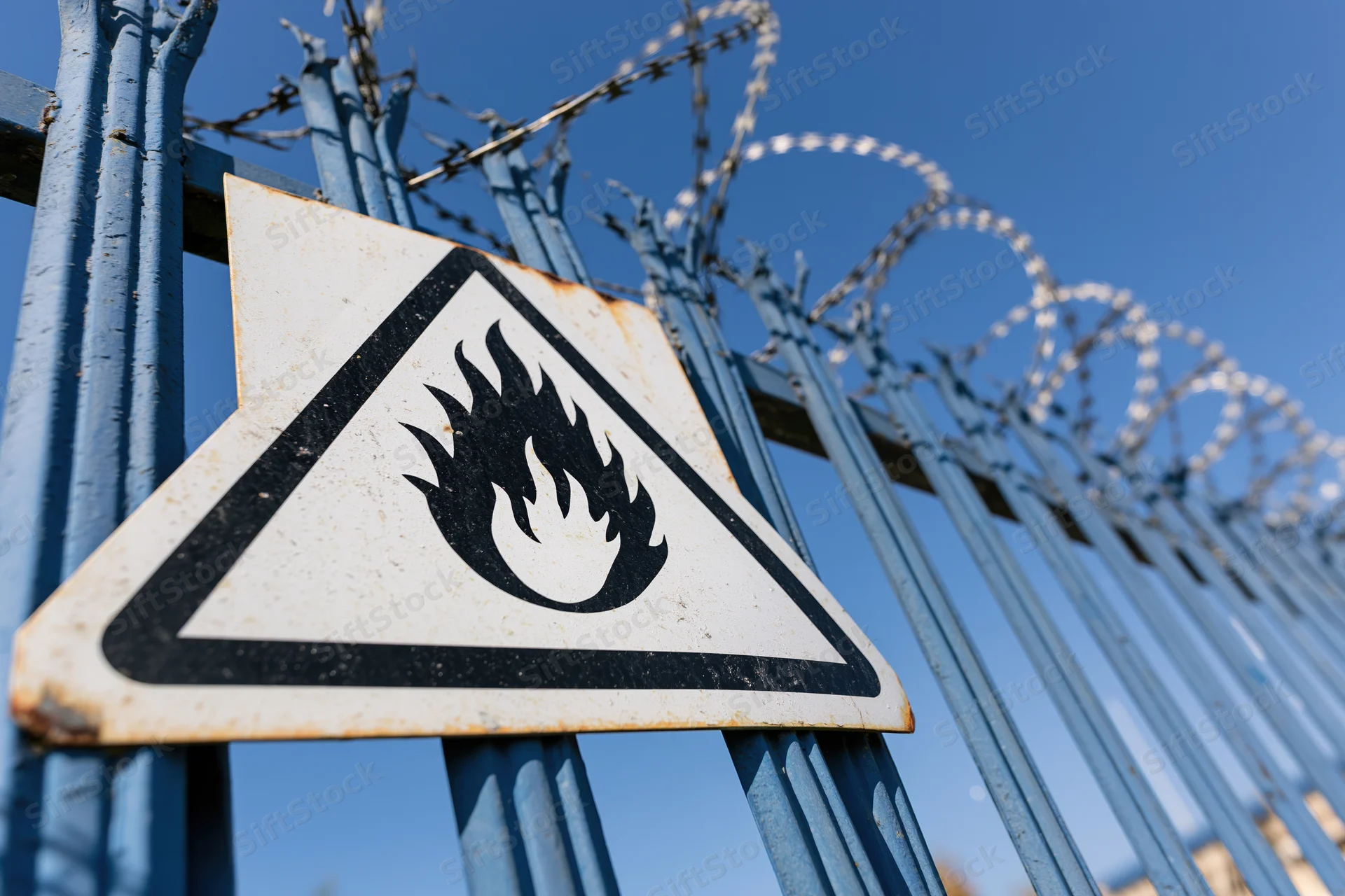 Triangular flammable warning sign mounted on blue metal fence with barbed wire against clear blue sky