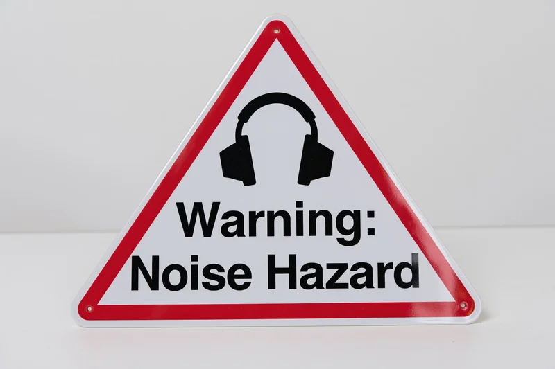 Triangular warning sign with red border and headphones symbol indicating noise hazard on white background