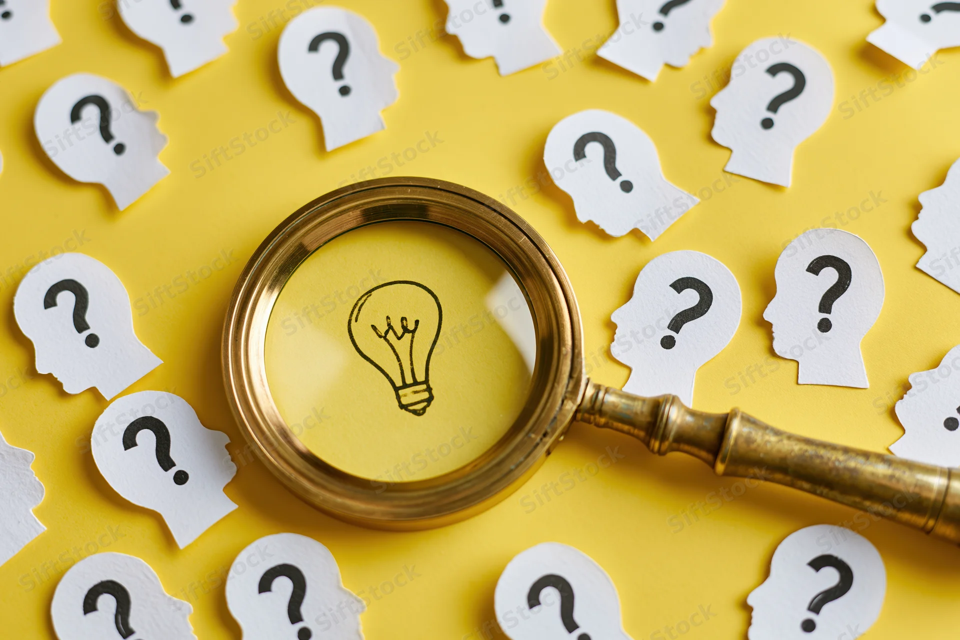 Vintage brass magnifying glass over paper cutouts of heads with question marks on yellow background, focusing on a drawn lightbulb symbolizing idea discovery