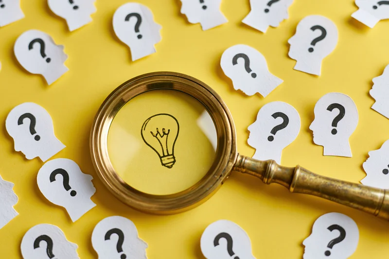 Vintage brass magnifying glass over paper cutouts of heads with question marks on yellow background, focusing on a drawn lightbulb symbolizing idea discovery
