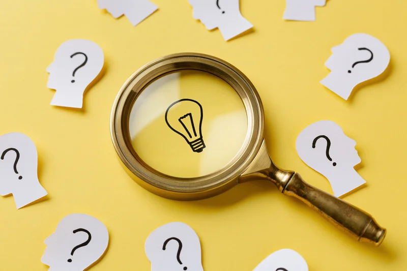 Vintage brass magnifying glass over a lightbulb drawing on yellow surface surrounded by paper head silhouettes with question marks
