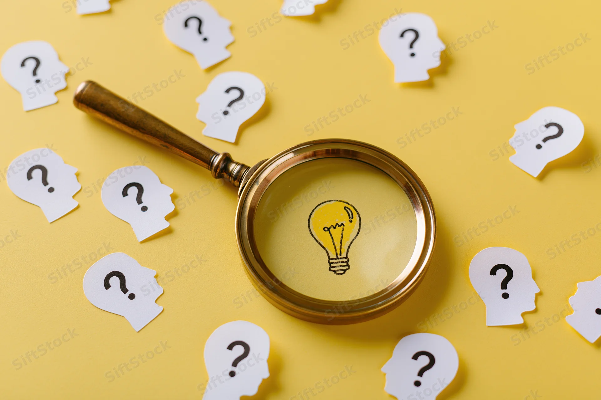 Vintage brass magnifying glass over yellow surface with paper head cutouts containing question marks and a lightbulb visible through the lens