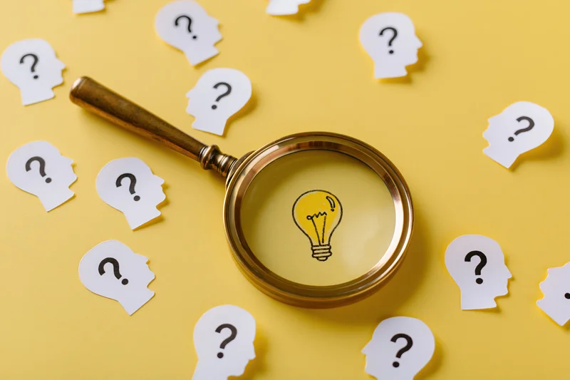 Vintage brass magnifying glass over yellow surface with paper head cutouts containing question marks and a lightbulb visible through the lens