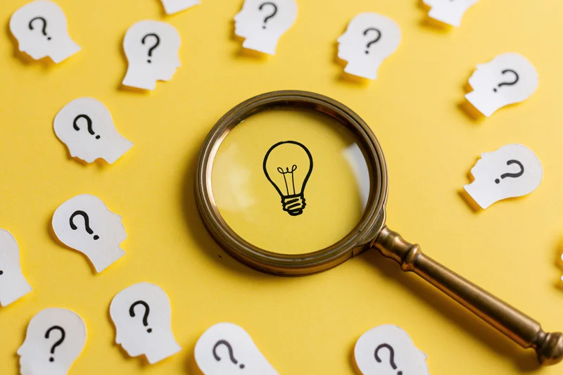 Vintage brass magnifying glass focusing on a lightbulb drawing among paper head cutouts with question marks on a yellow surface