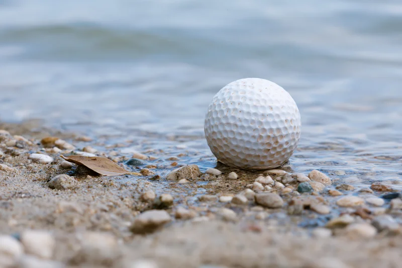 White golf ball resting on wet sandy shore among small pebbles near water