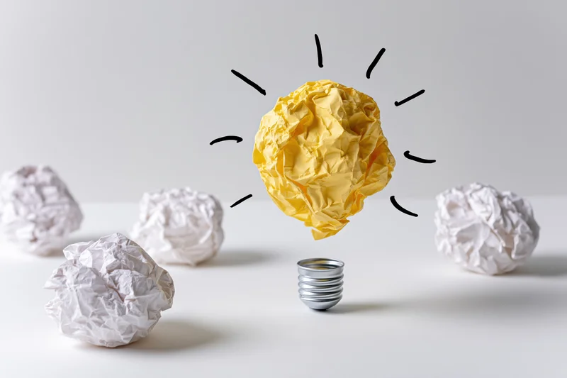 Yellow crumpled paper ball shaped as glowing lightbulb surrounded by white paper balls on white surface