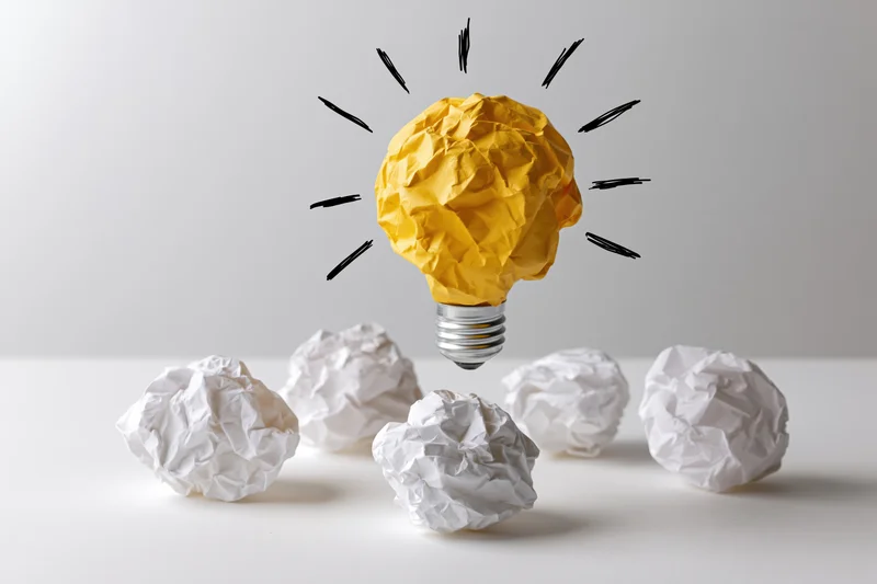 Yellow crumpled paper ball shaped as glowing lightbulb floating above white paper balls on plain surface