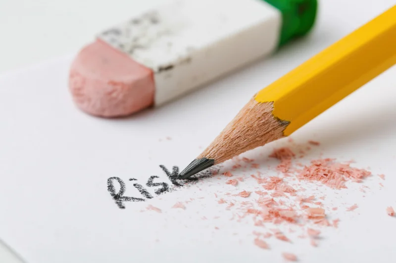 Yellow pencil erasing the word risk on white paper with pink eraser debris nearby