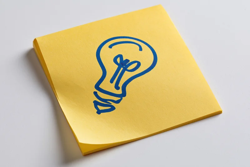 Yellow sticky note with hand-drawn blue lightbulb on white surface