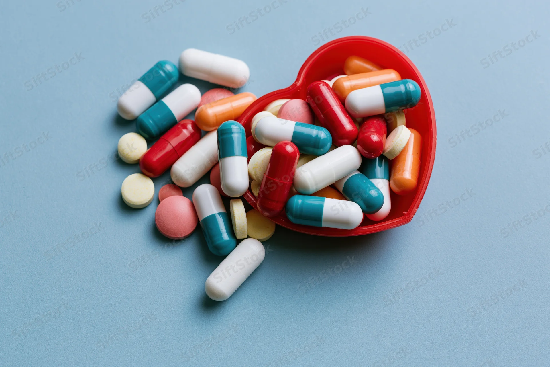 Assorted pharmaceutical capsules and tablets spilling from a red heart-shaped container on a blue surface