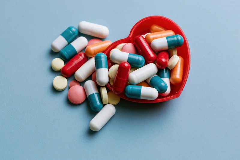 Assorted pharmaceutical capsules and tablets spilling from a red heart-shaped container on a blue surface